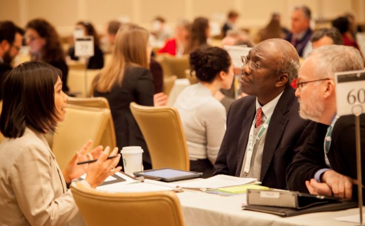 Two men in suits interview a woman at a table in a conference