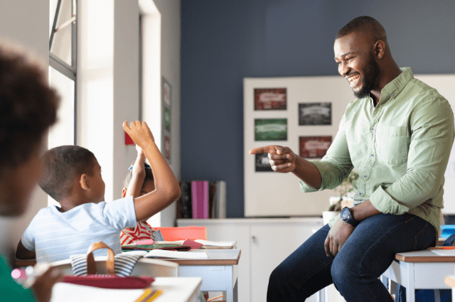 African American male teacher sitting casually on a desk pointing with a smile to a student