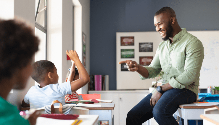 African American male teacher sitting casually on a desk pointing with a smile to a student