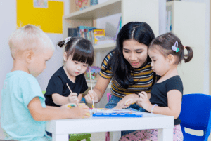 A young Asian female teacher helps three very young students paint on paper