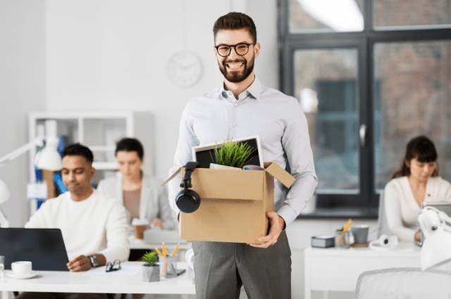 smiling young male businessman in an office holding a box of his desk items that he has packed up