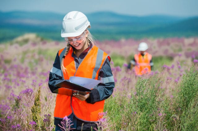 Female student in wildflower field with hard hat and writing down observations on a clipboard