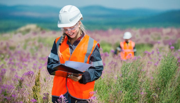 Female student in wildflower field with hard hat and writing down observations on a clipboard