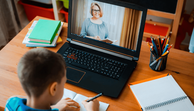 Female teacher on laptop screen smiling while young boy writes notes