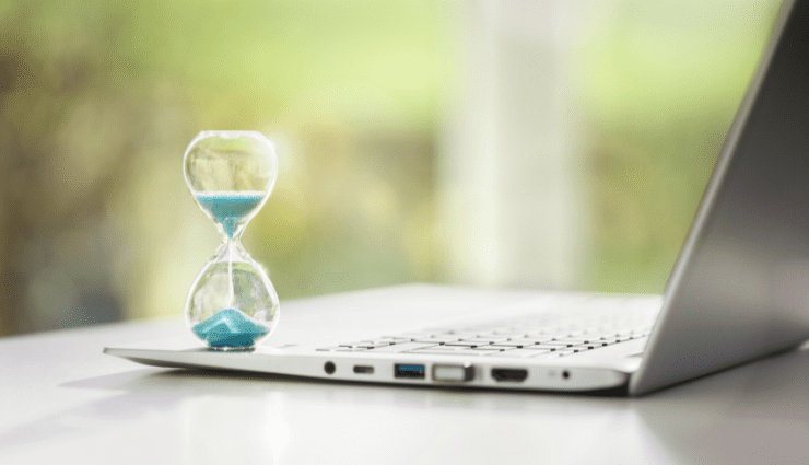 Hourglass with teal sand sitting next to an open laptop computer