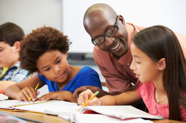 Teacher Helping Pupils Studying At Desks In Classroom