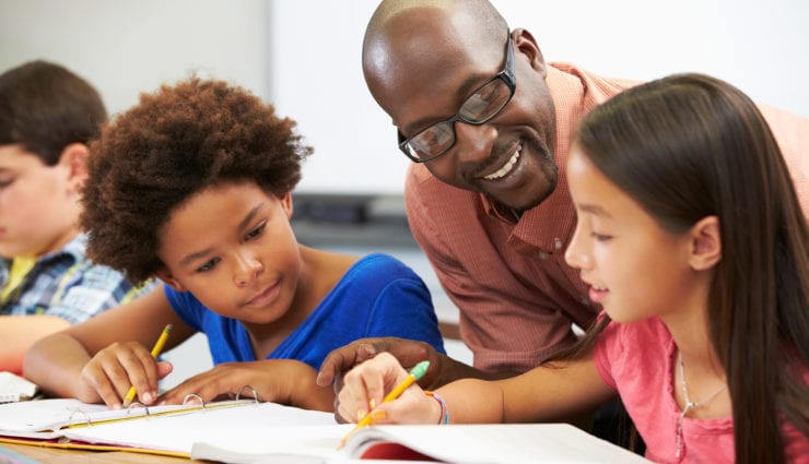 Teacher Helping Pupils Studying At Desks In Classroom