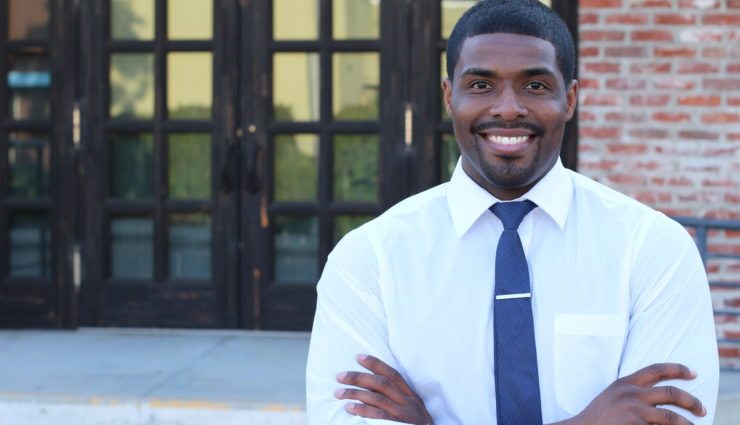 Male teacher with his arms crossed smiling in front of a school door outside