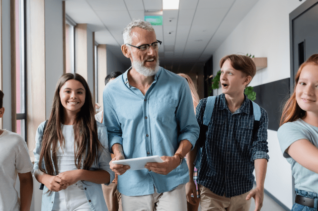 smiling male teacher with gray beard walking through hallway with four smiling middle school students