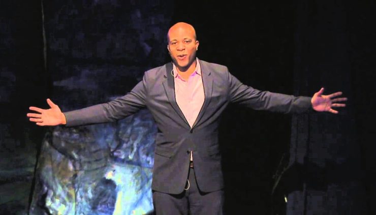 Derrick Gay speaks on stage with arms open