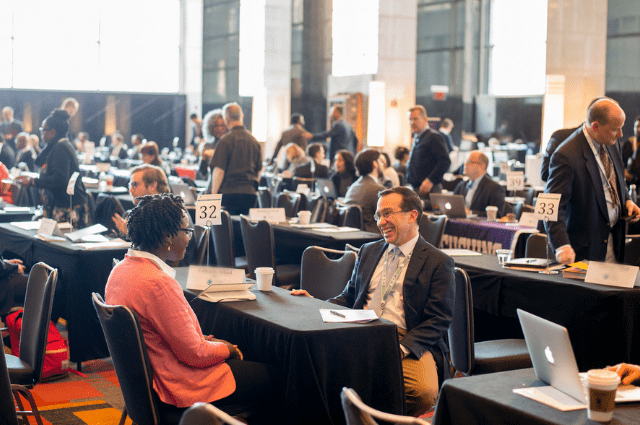 A large ballroom of tables and attendees with a focus on one table with a man and woman smiling and speaking