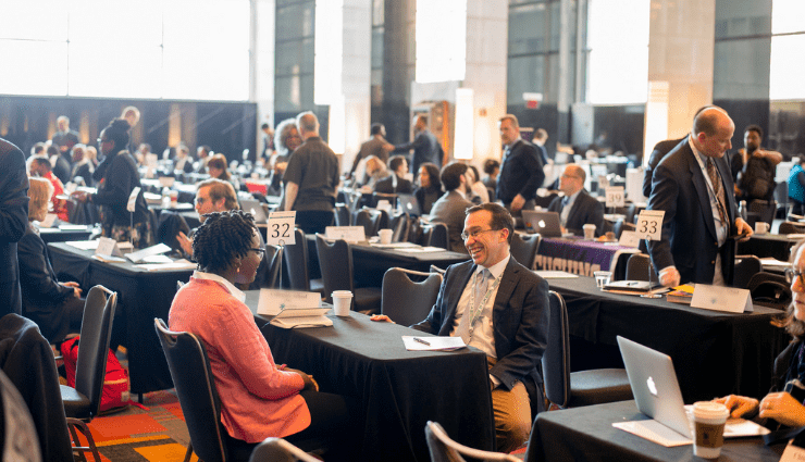 A large ballroom of tables and attendees with a focus on one table with a man and woman smiling and speaking