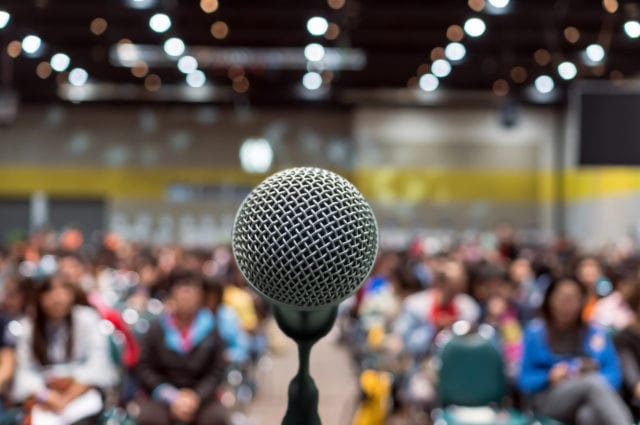 Microphone over photo of conference hall or seminar room in Exhibition Center background with Speakers on the stage and attendee background