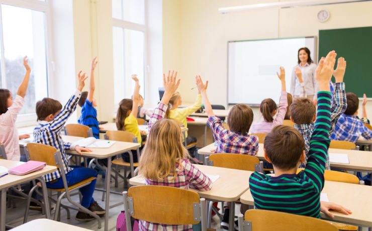 Teacher stands in front of class of elementary age students with raised hands