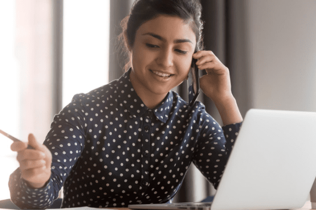 woman talking on phone with a laptop and taking notes on paper