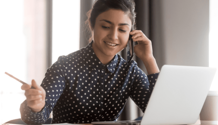 woman talking on phone with a laptop and taking notes on paper