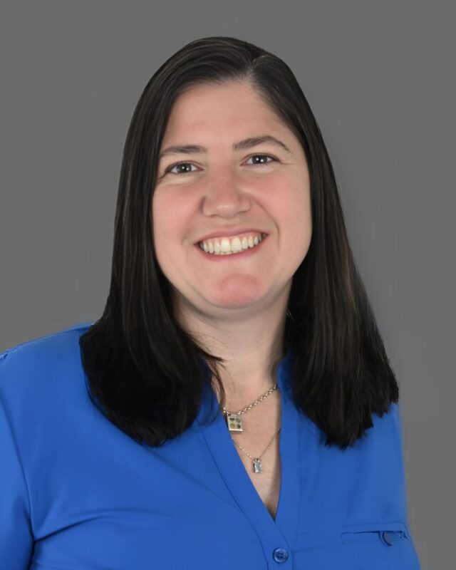 Smiling person with long dark hair wearing a blue shirt on a gray background.