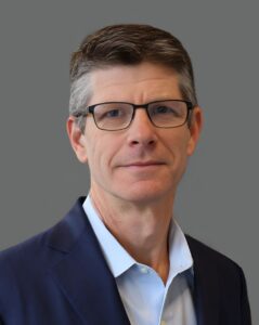 Man with glasses and short hair in a dark suit against a gray background.