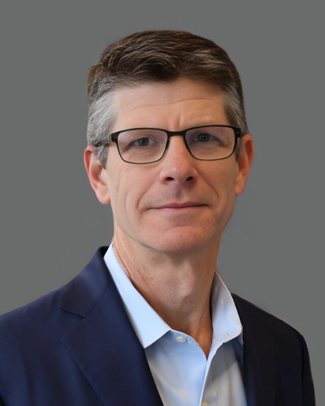 Man with glasses and short hair in a dark suit against a gray background.