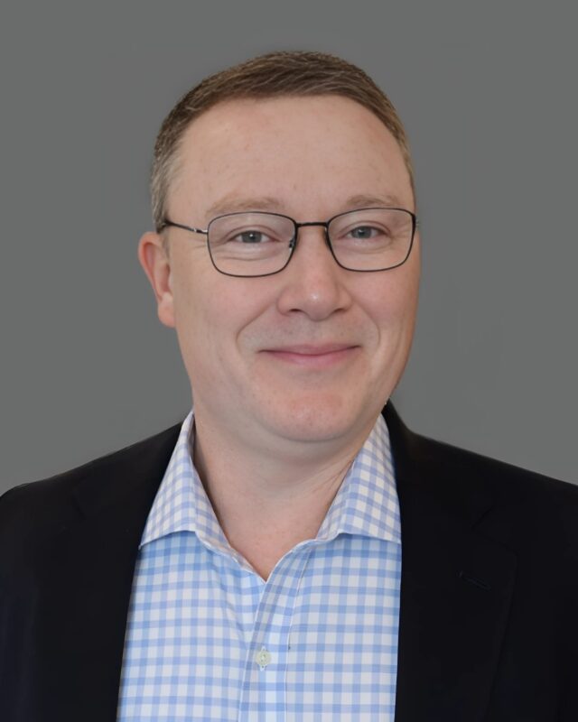 Smiling man in glasses and suit jacket with a light blue checkered shirt against a gray background.