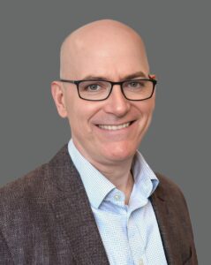 Smiling bald man wearing glasses and a brown jacket against a gray background.