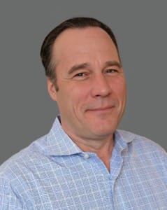 Man in a light blue checkered shirt against a gray background, smiling slightly.