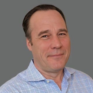 Man in a light blue checkered shirt against a gray background, smiling slightly.