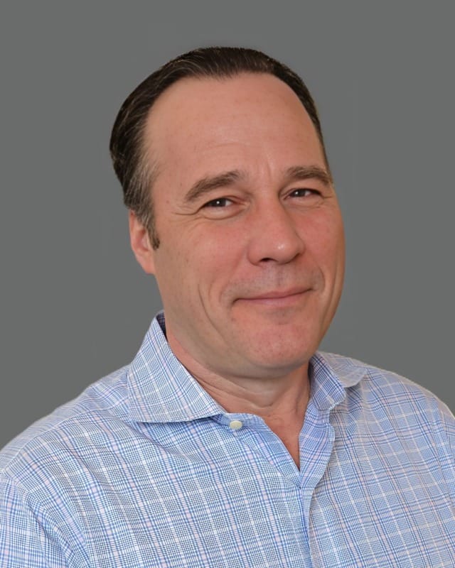 Man in a light blue checkered shirt against a gray background, smiling slightly.