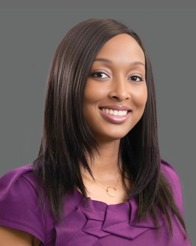 Smiling person with long hair wearing a purple blouse against a gray background.