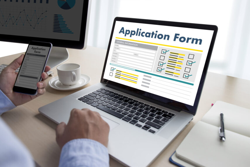 Person filling out an online application form on a laptop, with a smartphone displaying the same form.