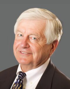 Older man in a suit and striped tie smiling against a gray background.