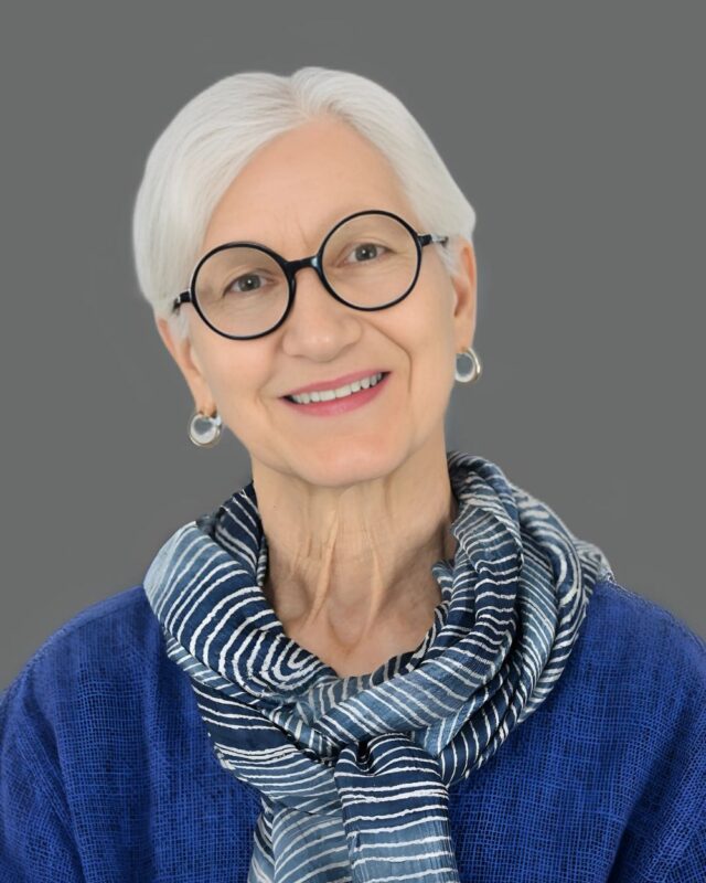 Elderly woman with white hair and glasses, wearing a blue scarf and sweater, smiling against a gray background.