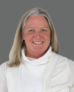 Smiling person with long gray hair wearing a white turtleneck against a gray background.