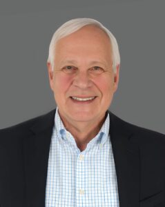 Smiling man with gray hair in a dark suit and checkered shirt against a gray background.
