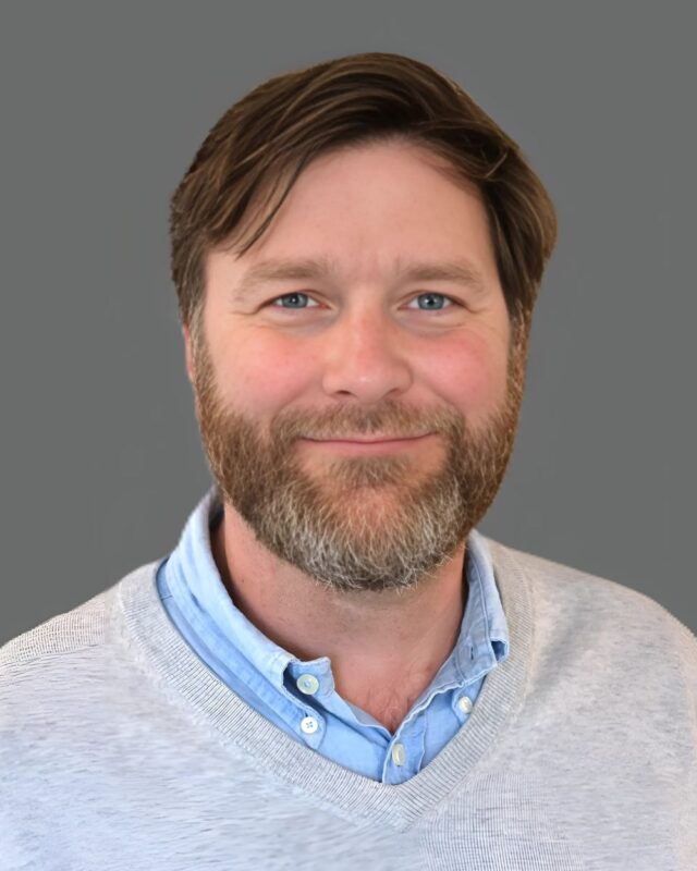 Man with a beard and short hair smiling, wearing a light blue shirt and gray sweater, against a gray background.