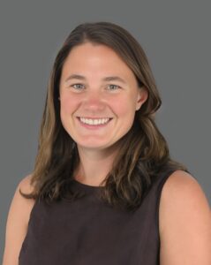 Smiling person with long brown hair, wearing a black top against a gray background.