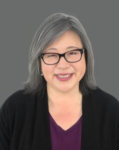 Smiling person with glasses and gray hair against a gray background.