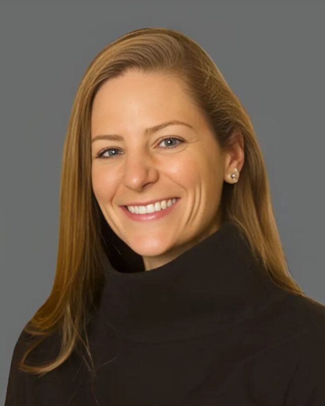 Smiling woman with long hair wearing a black turtleneck against a grey background.