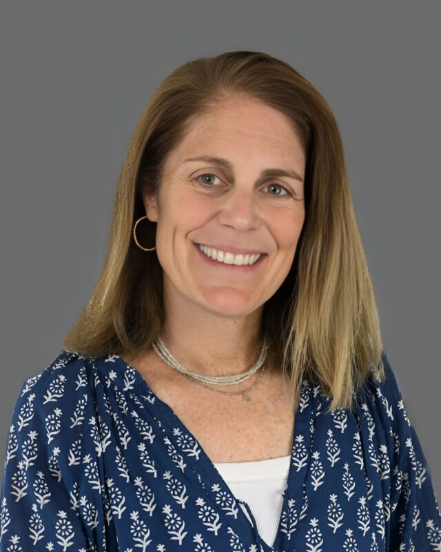 Smiling person with shoulder-length hair wearing a blue patterned blouse and necklace against a gray background