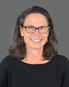 Smiling person with glasses and dark hair wearing a black top against a gray background.