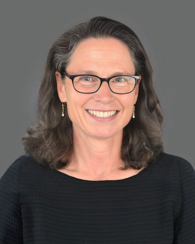 Smiling person with glasses and dark hair wearing a black top against a gray background.