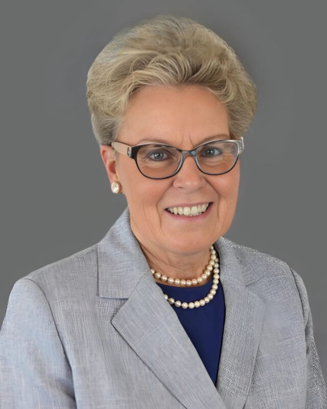 Smiling person with glasses, wearing a gray blazer and pearl necklace, against a gray background.