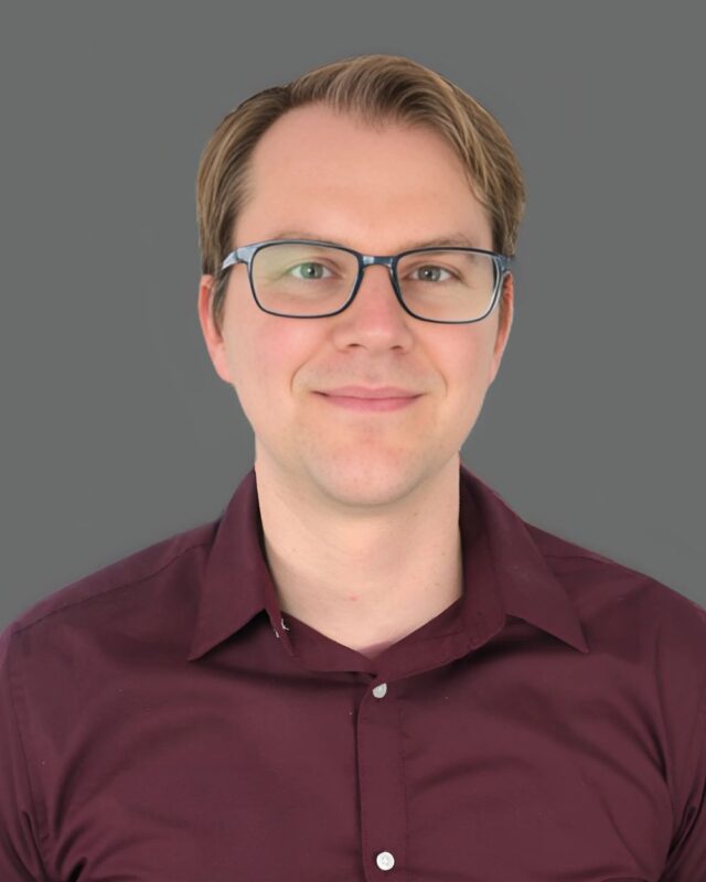 Smiling person with glasses and wearing a maroon shirt against a gray background.