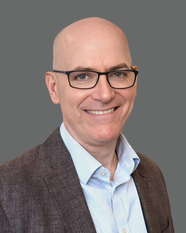 Bald smiling man wearing glasses and a brown jacket against a gray background.