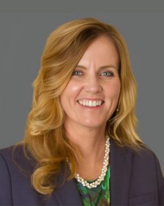Smiling person with long blonde hair wearing a dark blazer over a green shirt and necklace against a gray background.