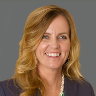 Smiling person with long blonde hair wearing a dark blazer over a green shirt and necklace against a gray background.
