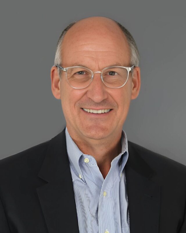 Bald man with glasses wearing a suit and tie, smiling against a gray background.