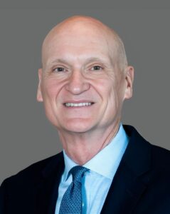 Bald man in a dark suit with a light blue shirt and tie, smiling against a gray background.