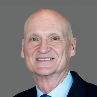 Bald man in a dark suit with a light blue shirt and tie, smiling against a gray background.