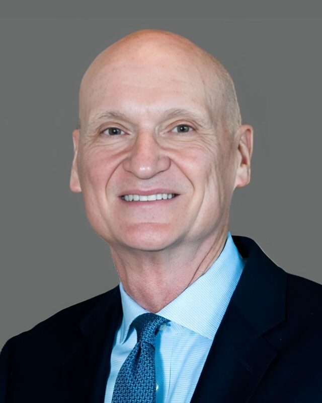 Bald man in a dark suit with a light blue shirt and tie, smiling against a gray background.